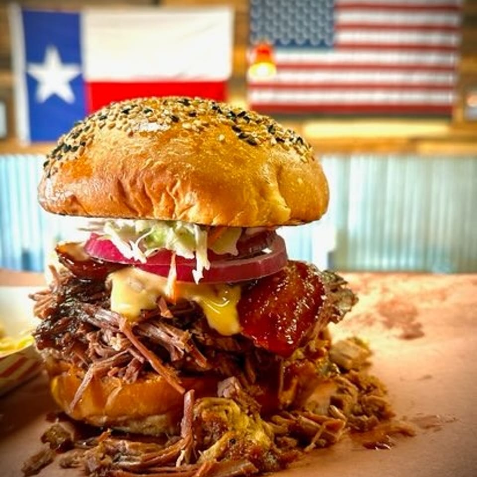 Loaded barbecue pulled-pork burger with melted cheese, coleslaw, onions, and sesame bun on a wooden counter.